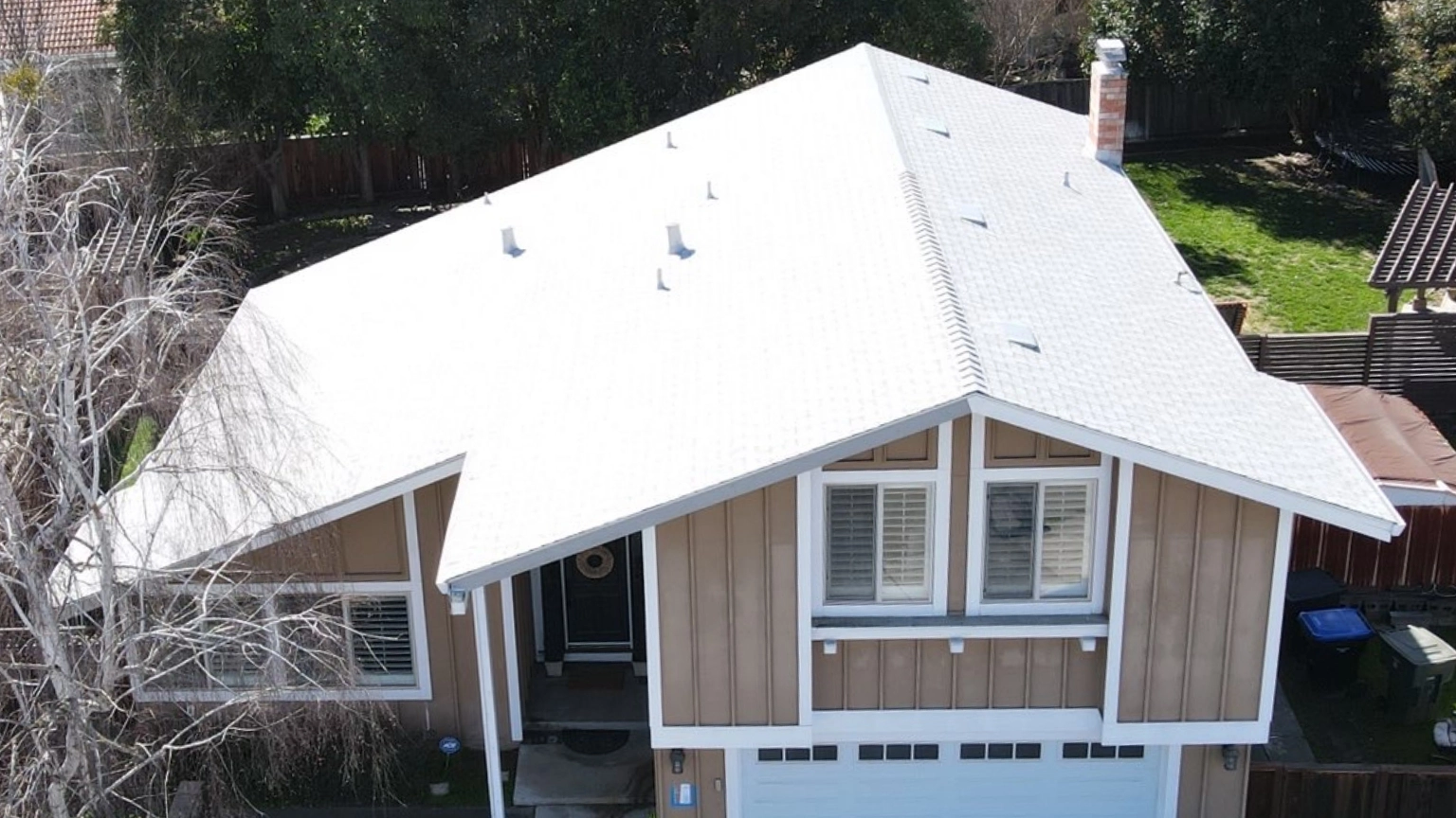 A two-story house with a white roof, beige and white exterior, large front windows, and a white garage door, surrounded by a fenced yard and trees.