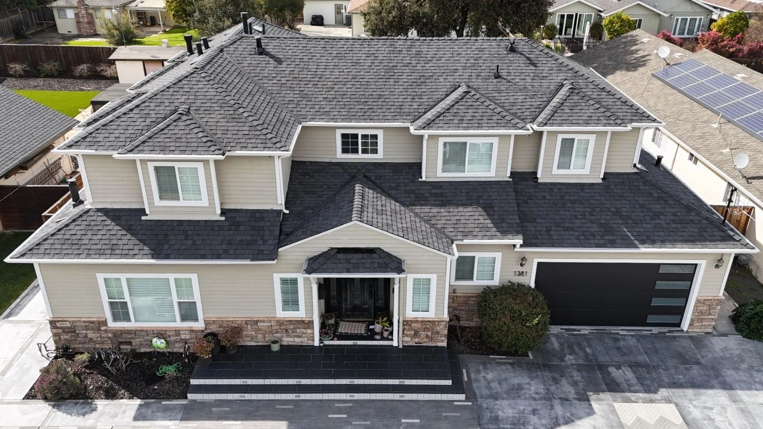 A two-story suburban house with beige siding, stone accents, dark roof shingles, and a black double garage, photographed from above.