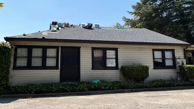 A single-story house with beige siding, black trim, and several items scattered on the roof, seen from the street during the day.