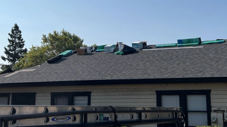 Several bundles of roofing materials and boxes are stacked on the roof of a single-story house, with trees and clear sky in the background.