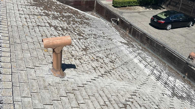 A shingled roof with patches of missing granules, a brown vent pipe, and a parking lot with cars in the background.