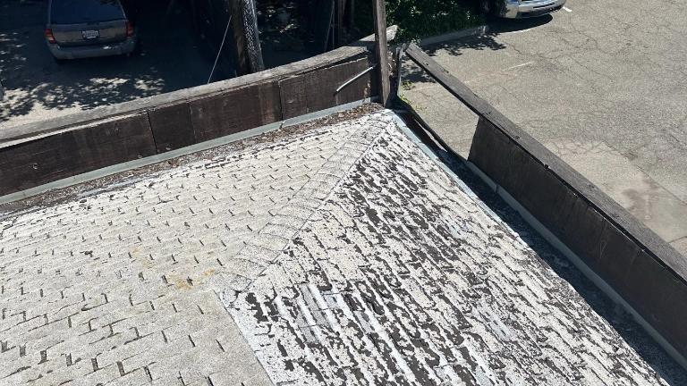 View of a weathered, shingle-covered rooftop with peeling paint, surrounded by a wooden railing and overlooking a parking lot with cars.