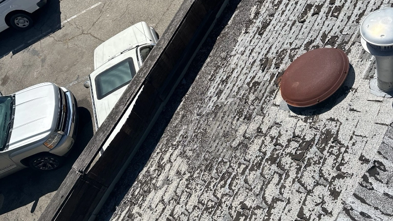 View from above of a weathered, partially white rooftop with a round vent and metal pipe, next to a parking lot with two white vehicles.