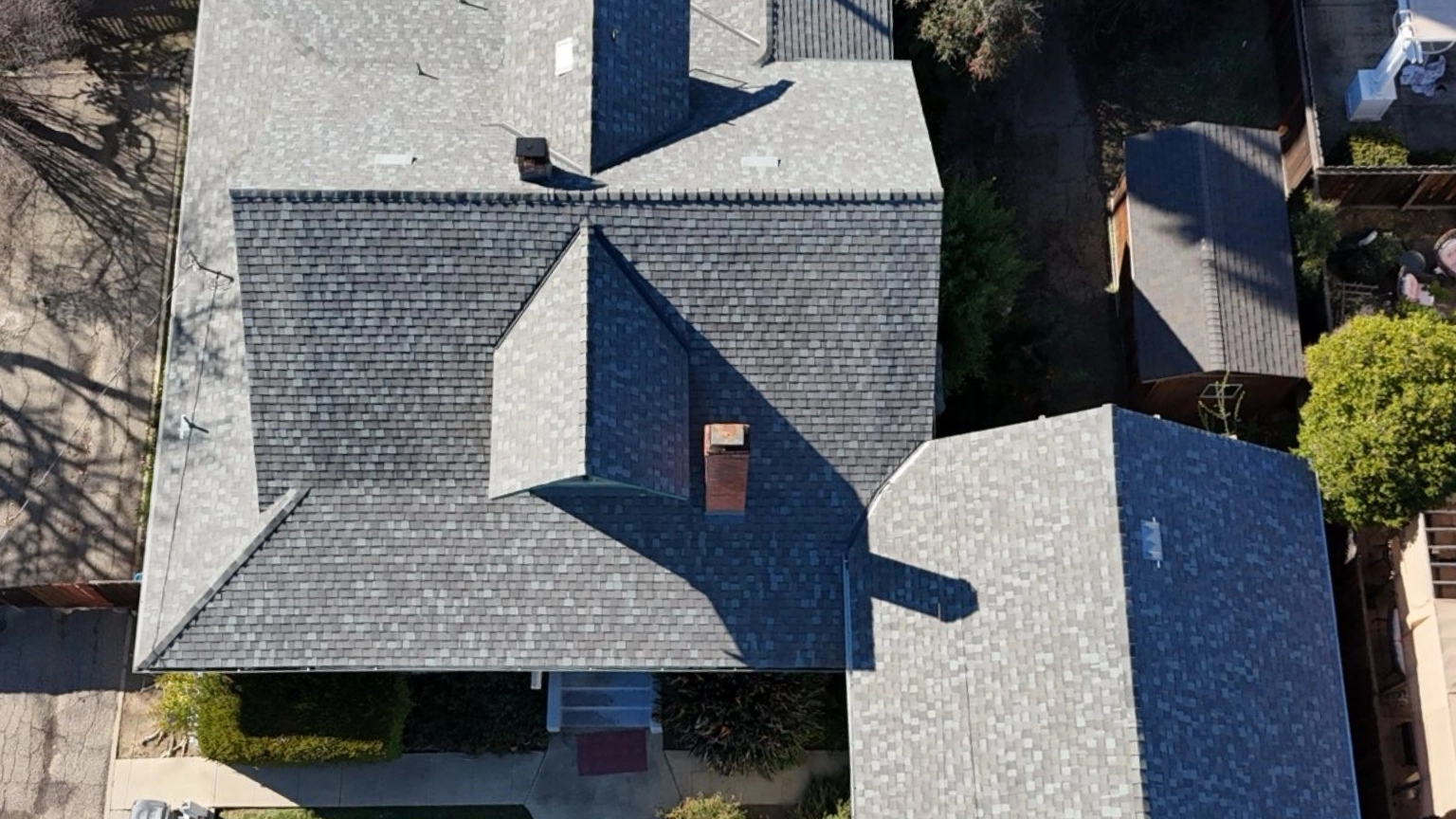 Aerial view of two gray-shingled rooftops with intersecting rooflines, surrounded by trees, fencing, and a concrete walkway.