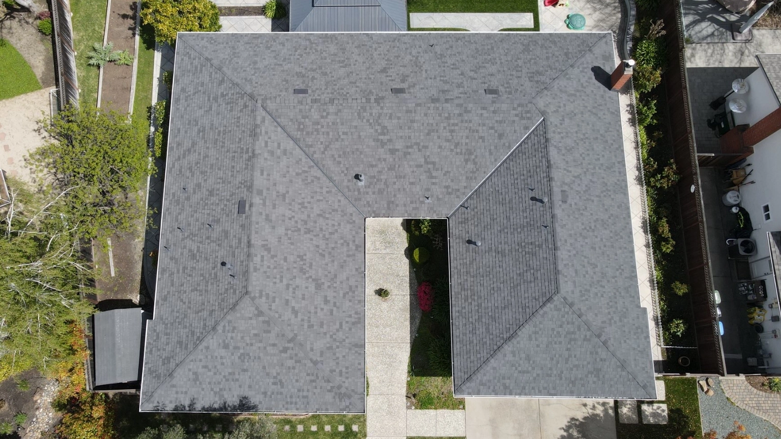 Aerial view of a gray-shingled, L-shaped residential roof surrounded by trees, pathways, and landscaped yards.