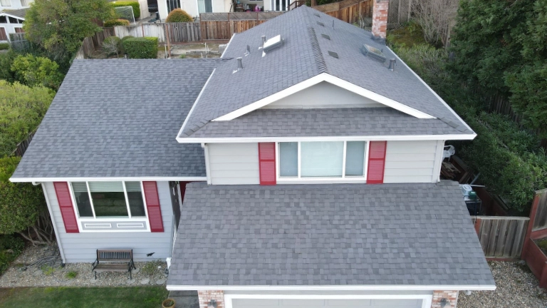 Aerial view of a two-story suburban house with gray siding, gray roof, white trim, and red shutters, surrounded by trees and a fenced backyard.