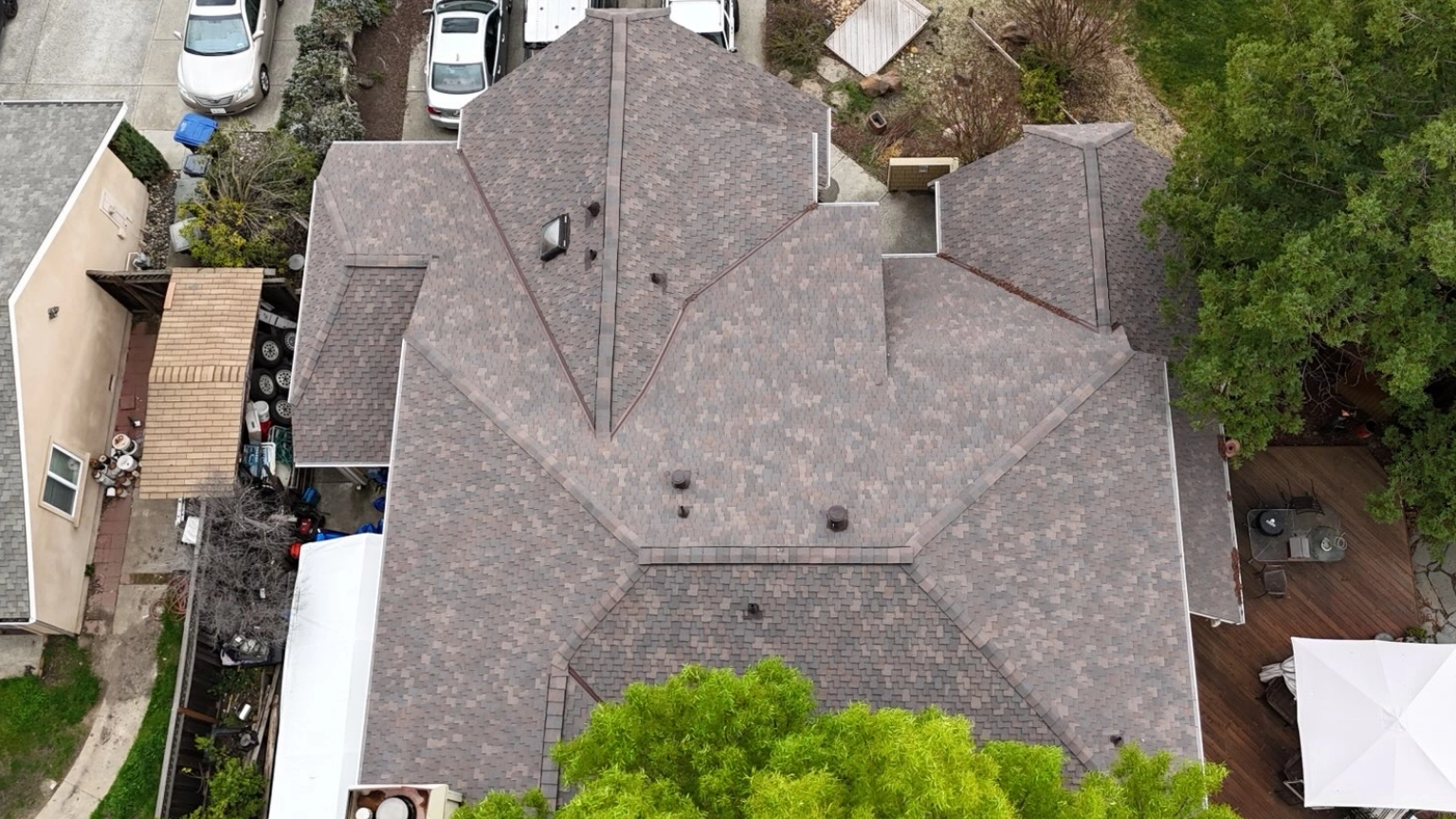 Aerial view of a residential house with a complex gray shingle roof, surrounded by trees, parked cars, and neighboring buildings.
