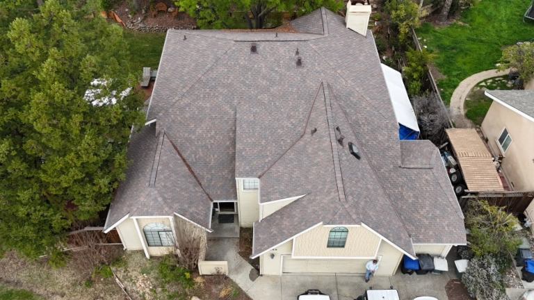 Aerial view of a beige two-story house with a gray shingle roof, surrounded by trees and greenery, with a driveway and person standing near the entrance.