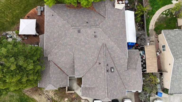 Aerial view of a house roof with gray shingles, adjoining backyard, patio deck with a canopy, trees, and neighboring houses visible.