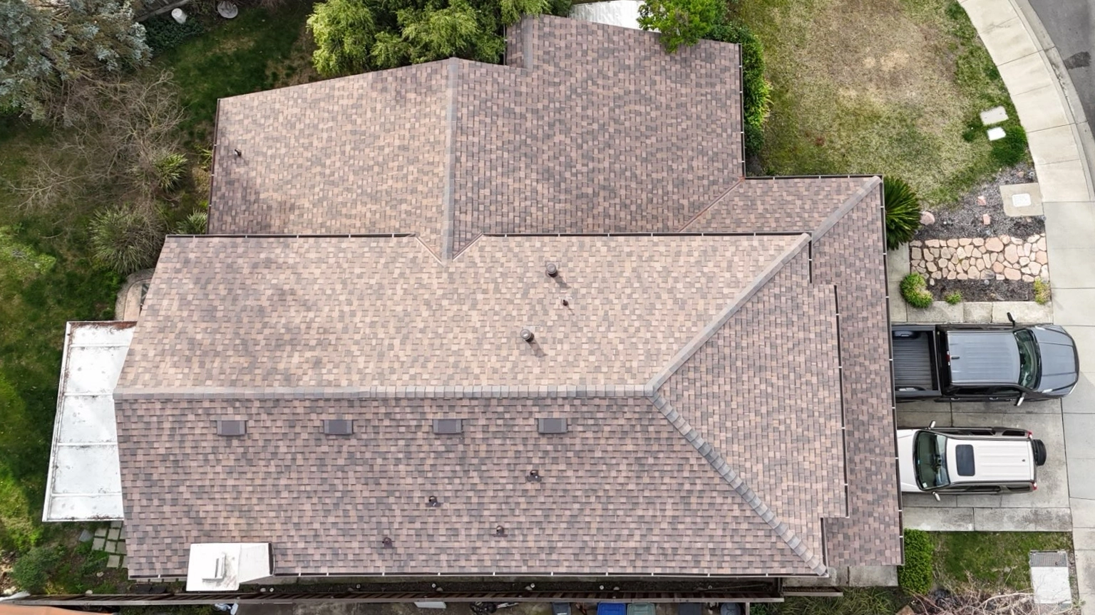 Aerial view of a house with a brown shingle roof, two vehicles parked in the driveway, and a landscaped yard with trees and grass.
