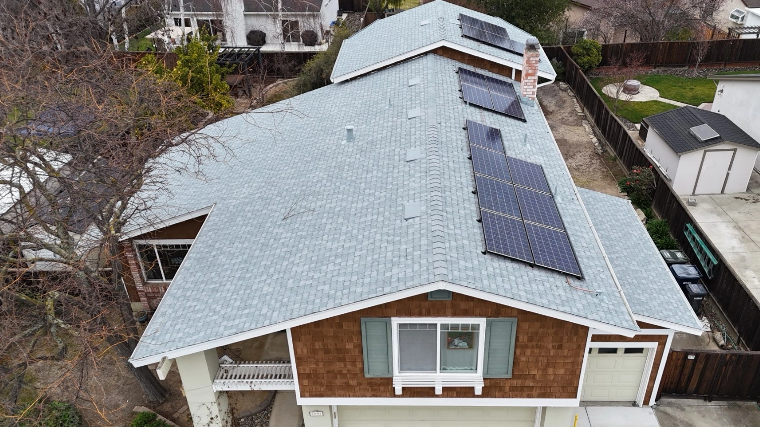 Aerial view of a house with light gray shingles and solar panels installed on the roof, surrounded by trees and neighboring homes.