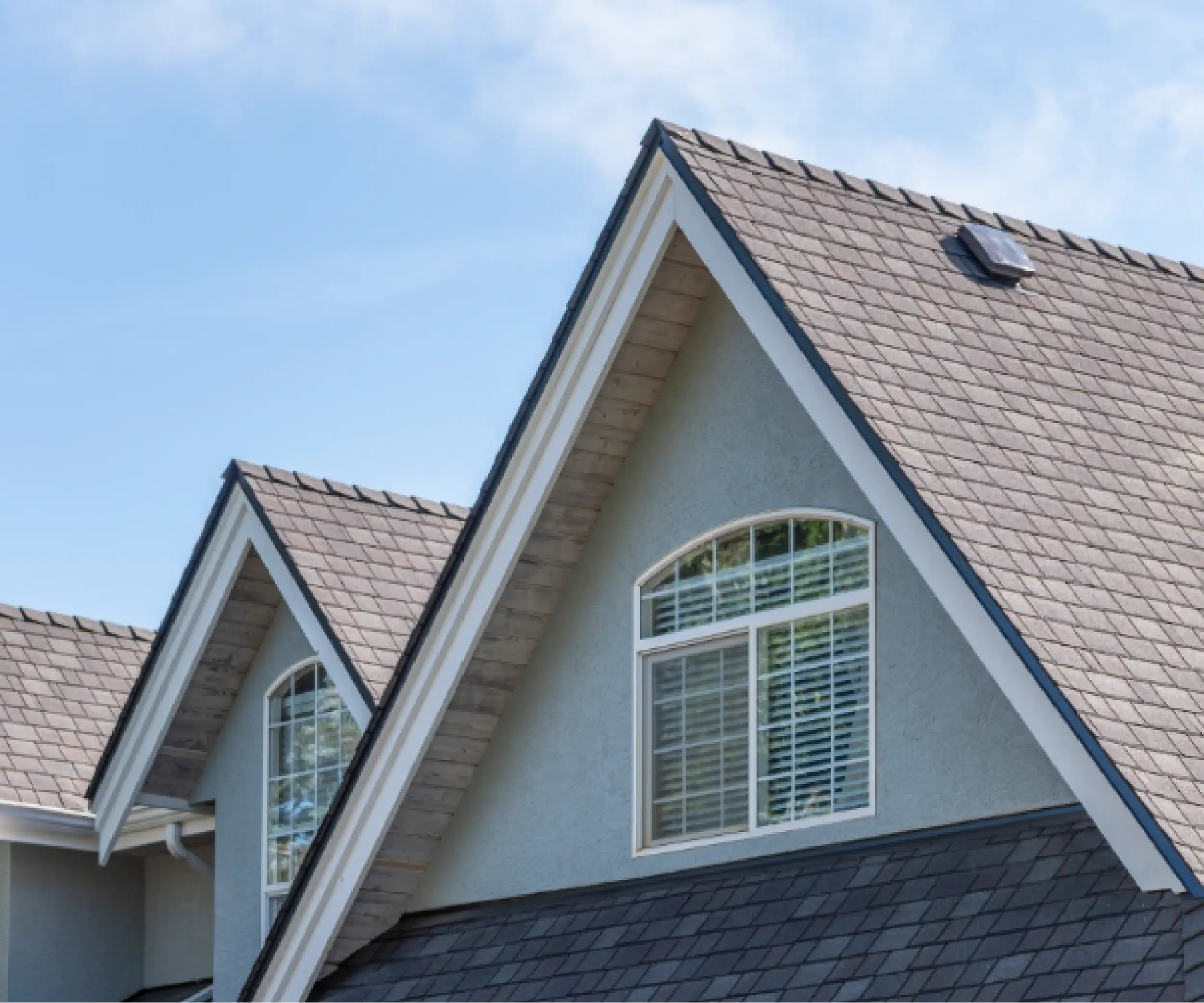 Close-up view of a house roof with gray shingles, light blue exterior walls, and two arched windows with white trim and blinds—showcasing quality residential roofing in Blackhawk under a clear blue sky.