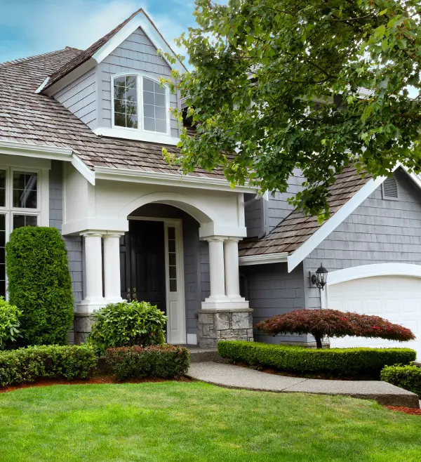 Gray two-story suburban house with white trim, arched entryway, attached garage, landscaped lawn, shrubs, and a large tree in the front yard—recently updated with expert dry rot & structural repair for lasting durability.