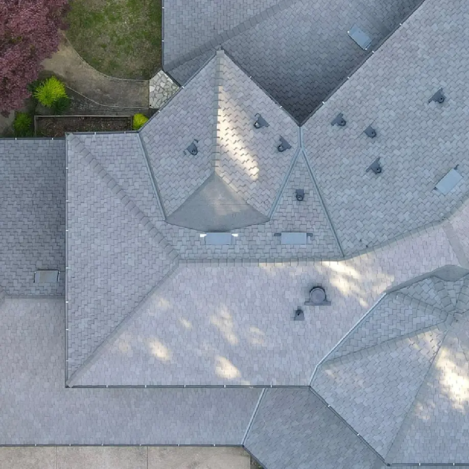 Aerial view of a gray shingled residential roofing system with multiple peaks and vents, bordered by some trees and a small section of yard.
