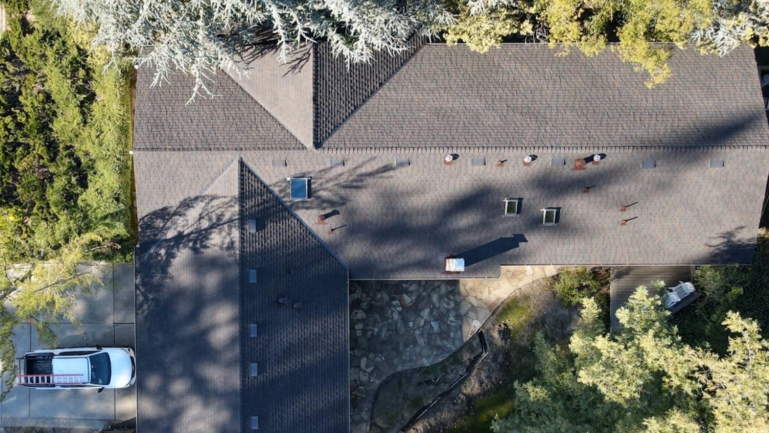 Aerial view of a house roof with several vents and skylights, surrounded by trees. Evidence of recent re-roofing in Alamo is visible, and a white vehicle is parked in the driveway on the left.