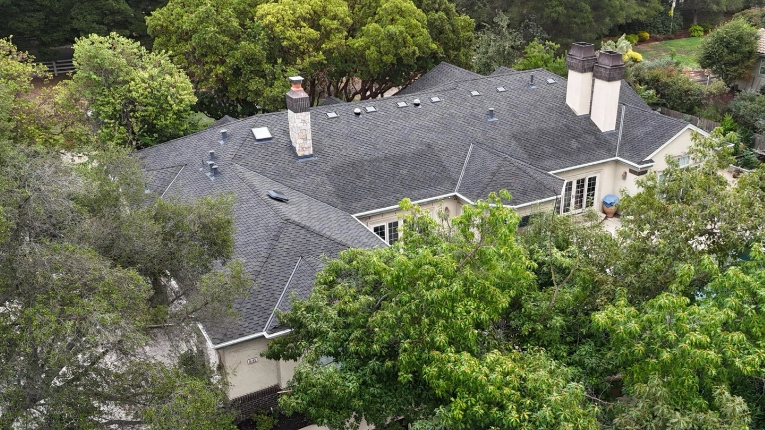 Aerial view of a large house with a dark gray shingle roof, multiple chimneys, and new gutters after re-roofing in Pleasanton, all surrounded by dense green trees and foliage.