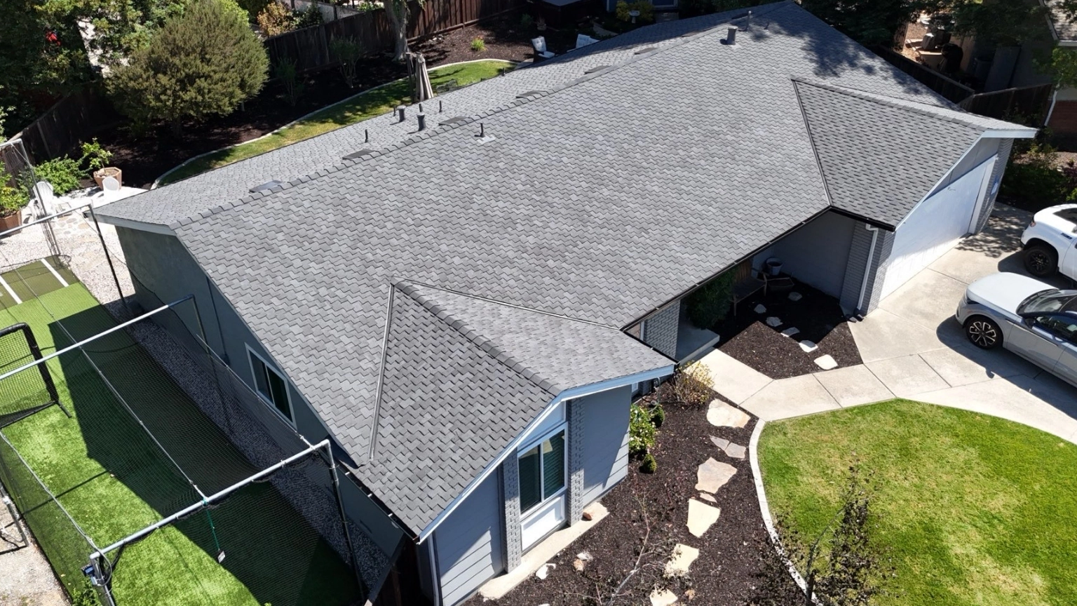 Aerial view of a single-story gray house with a shingled roof, green lawn, stone pathway, and a white car parked in the driveway—showcasing expert re-roofing with new gutters and downspouts in San Ramon.